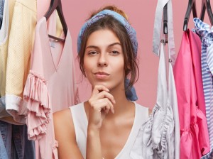 Portrait of thoughtful woman with scarf on head, standing near hangers with clothes, thinking over what to buy. Female shopaholic in boutique with pensive expression, having difficult choice